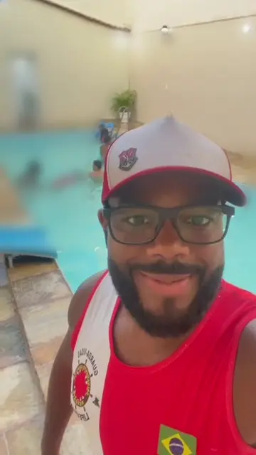 O guardião da Oliveira Piscinas, de boné e regata vermelha, na beira da piscina de um salão de festas, com as crianças desfocadas ao fundo